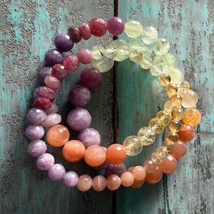 May include: Three layered bracelets made of semi-precious stones. The top layer is pink and red, the middle layer is purple, and the bottom layer is orange.