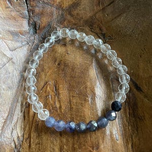 May include: A beaded bracelet featuring clear, faceted beads and a section of blue-purple beads. A single black bead is also included in the design. The bracelet is displayed on a wooden surface.