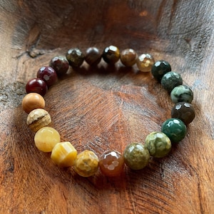 May include: A beaded bracelet featuring a variety of colorful, faceted gemstone beads. The beads range in color from deep red and brown to yellow and green. The bracelet is displayed on a wooden surface.