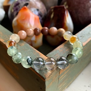 May include: A beaded bracelet featuring a variety of natural gemstones. The bracelet includes green, clear, and brown beads, with a mix of colors and textures. The bracelet is displayed on a distressed, light green wooden tray, with other decorative objects in the background.