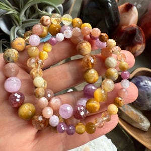 May include: Three beaded bracelets in shades of yellow, pink, and purple. The bracelets are made of various gemstones, including faceted and round beads. The bracelets are held in a hand, with other crystals and plants in the background.