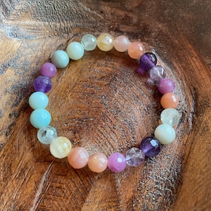 May include: A beaded bracelet featuring a variety of colorful gemstones. The bracelet includes round beads in shades of blue, purple, yellow, and peach. The bracelet is displayed on a wooden surface.