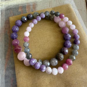 May include: Two strands of gemstone beads in shades of pink, purple, and gray. The beads are faceted and have a polished finish. The beads are arranged in a circle and are displayed on a brown fabric background.