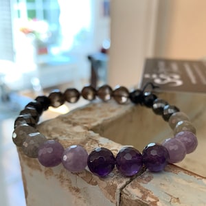 May include: A beaded bracelet featuring a gradient of colors. The bracelet transitions from dark gray to light gray, then to purple and amethyst-colored beads. The beads have a faceted cut, and the bracelet is strung on a black cord.