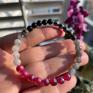 May include: A bracelet made with black, gray, and red gemstone beads. The bracelet is made with a variety of different types of stones, including black onyx, hematite, clear quartz, labradorite, and red ruby.