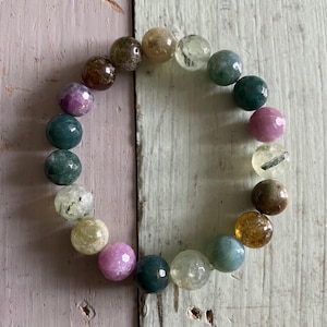 May include: A bracelet made of various semi-precious stones, including green, brown, pink, and clear stones. The bracelet is made of round beads and is strung on a clear elastic cord.