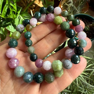 May include: A beaded bracelet featuring a variety of colorful gemstones. The bracelet includes green, pink, and white beads. The beads have a faceted cut, giving them a sparkling appearance. The bracelet is displayed against a backdrop of green foliage.