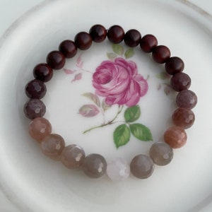 May include: A brown wooden beaded bracelet with a gradient of light pink, grey, and brown gemstone beads.