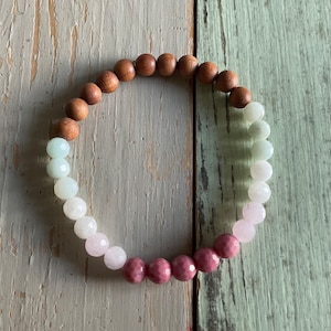 May include: A beaded bracelet featuring a mix of natural wood, white, pink, and dark pink beads. The bracelet is arranged in a circular shape, with the beads varying in size and color, creating a gradient effect. The bracelet is displayed on a light green wooden surface.