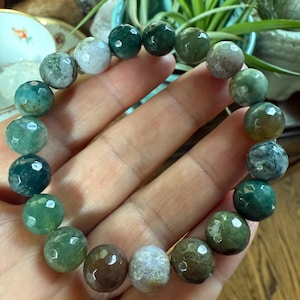 May include: A beaded bracelet featuring faceted, round gemstones in shades of green, brown, and white. The bracelet is held in a hand, showcasing the variety of colors and textures of the stones. The bracelet is a piece of jewelry.