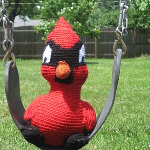 May include: A red crocheted cardinal bird toy sitting on a black swing set. The cardinal has black eyes, a black beak, and an orange nose.