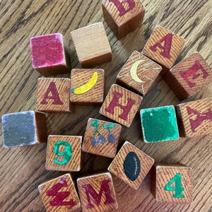 May include: A collection of wooden blocks featuring painted letters, numbers, and illustrations including a banana, a crescent moon, cherries, and a football. The blocks display various colors such as red, green, yellow, and black on natural wood surfaces.