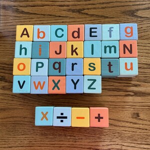 May include: A set of square wooden blocks featuring the alphabet in both uppercase and lowercase letters, along with four separate blocks displaying the mathematical symbols for multiplication, division, subtraction, and addition in various colors.