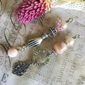 May include: A pair of dangle earrings featuring silver-tone metal components, including a hand charm and a fleur-de-lis charm. The earrings are accented with pink beads, cream-colored pearls, and a circular charm encrusted with clear rhinestones.