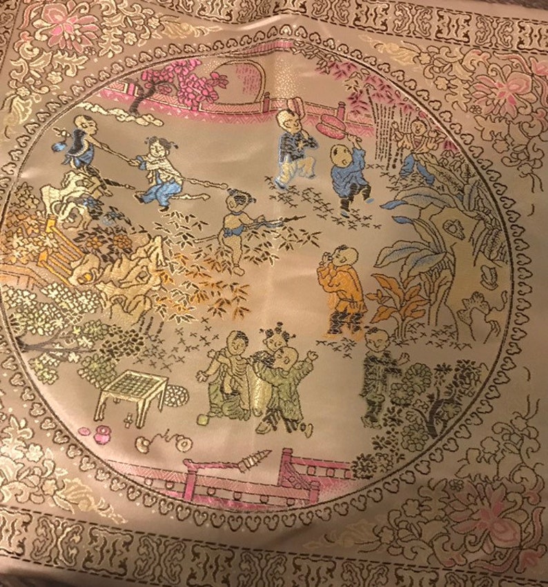 May include: A circular embroidered fabric panel with a beige background and a gold border. The panel features a scene of children playing in a garden with trees, flowers, and a bridge. The children are wearing traditional Chinese clothing.