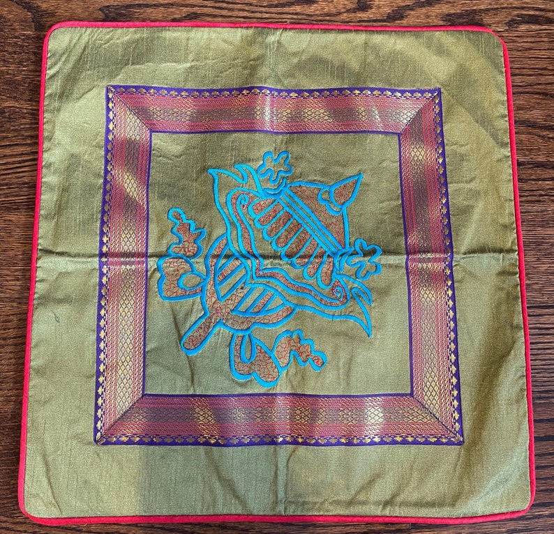 K&ouml;nnte beinhalten: Ein gr&uuml;nes Kissen mit rotem Rand zeigt ein blaues gesticktes Design eines stilisierten Vogels oder Tieres mit komplizierten Details. Das Design ist von einem goldenen und violetten Rand umrahmt.