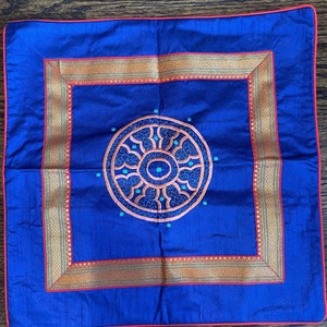 K&ouml;nnte beinhalten: Ein blaues Kissen mit einem goldenen und roten Stickrand und einem kreisf&ouml;rmigen Design in der Mitte. Das Design zeigt ein mandalaartiges Muster mit t&uuml;rkisfarbenen und orangefarbenen Akzenten.