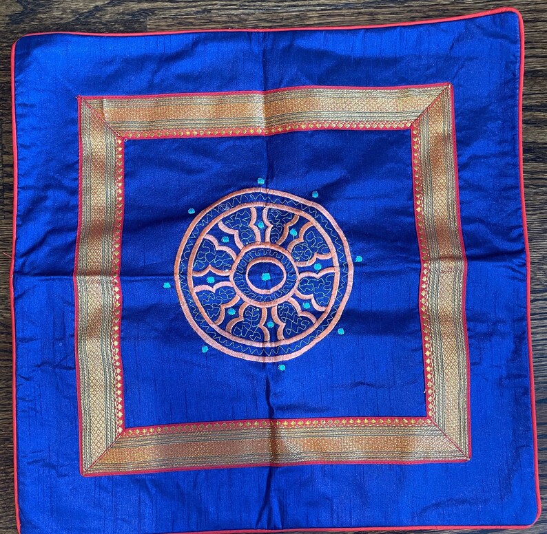 K&ouml;nnte beinhalten: Ein blaues Kissen mit einem goldenen und roten Stickrand und einem kreisf&ouml;rmigen Design in der Mitte. Das Design zeigt ein mandalaartiges Muster mit t&uuml;rkisfarbenen und orangefarbenen Akzenten.