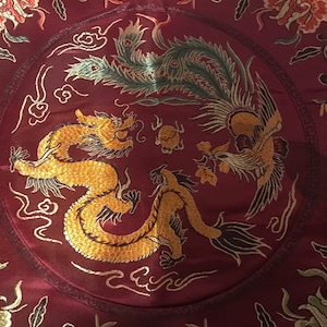 May include: A red silk cushion with a gold embroidered dragon and phoenix design. The cushion has a circular design with a border of gold and black.