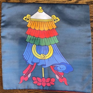 K&ouml;nnte beinhalten: Ein blaues Kissen mit einem farbenfrohen tibetischen Gebetsfahnen-Design. Die Fahne hat eine wei&szlig;e Spitze, gelbe, orange, rote, gr&uuml;ne und blaue Abschnitte. Die Fahne befindet sich auf einem blauen Hintergrund mit einer roten Lotusblume unten.