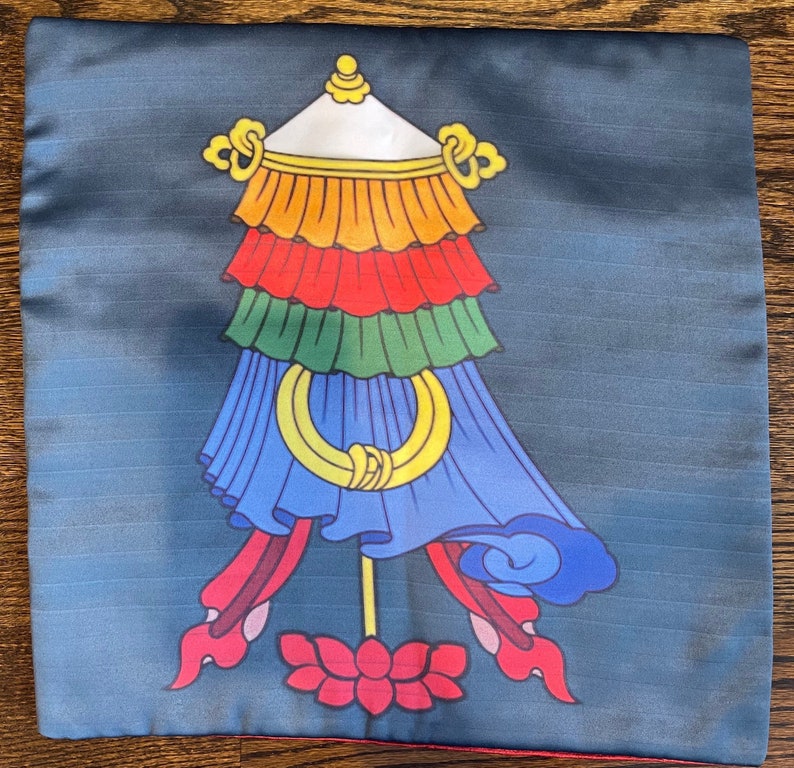 K&ouml;nnte beinhalten: Ein blaues Kissen mit einem farbenfrohen tibetischen Gebetsfahnen-Design. Die Fahne hat eine wei&szlig;e Spitze, gelbe, orange, rote, gr&uuml;ne und blaue Abschnitte. Die Fahne befindet sich auf einem blauen Hintergrund mit einer roten Lotusblume unten.