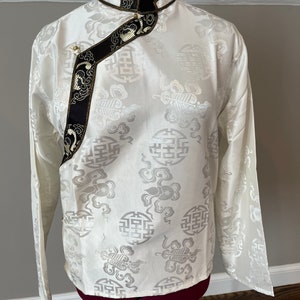 May include: A white silk shirt with a mandarin collar and long sleeves. The shirt has a black and gold trim and a pattern of stylized clouds and Chinese characters.