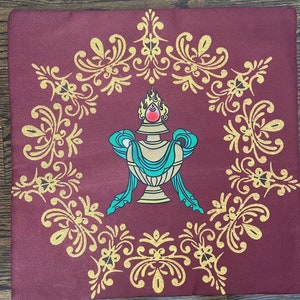 K&ouml;nnte beinhalten: Ein quadratisches Kissen aus burgunderfarbenem Stoff mit einem goldenen, verzierten Design und einem zentralen Bild einer goldenen Vase mit einem blaugr&uuml;nen Band und einem roten Juwel oben. Die Vase ist von einer goldenen Flamme umgeben.