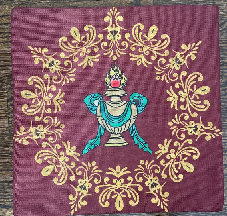 K&ouml;nnte beinhalten: Ein quadratisches Kissen aus burgunderfarbenem Stoff mit einem goldenen, verzierten Design und einem zentralen Bild einer goldenen Vase mit einem blaugr&uuml;nen Band und einem roten Juwel oben. Die Vase ist von einer goldenen Flamme umgeben.