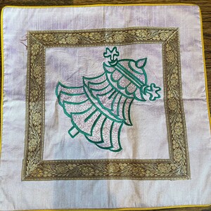 K&ouml;nnte beinhalten: Ein quadratisches Kissen mit helllila Hintergrund und goldener Stickerei an der Kante. In der Mitte des Kissens befindet sich ein gesticktes Design in Gr&uuml;n und Lila, das einen stilisierten Baum darstellt.