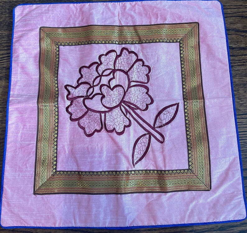 K&ouml;nnte beinhalten: Ein rosa Kissen mit einem goldenen und braunen bestickten Rand und einem rosa Blumendesign in der Mitte.