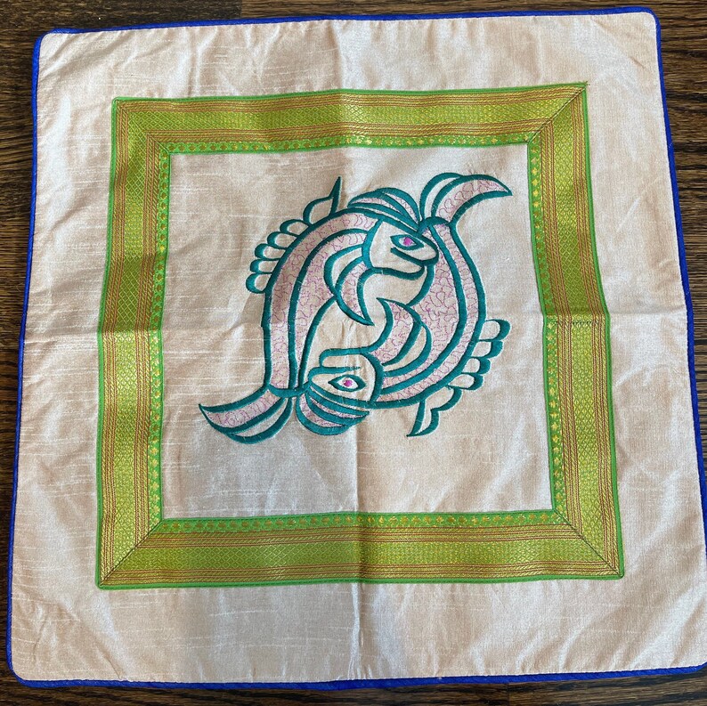 K&ouml;nnte beinhalten: Ein wei&szlig;es Kissen mit gr&uuml;nem und goldenem Rand. In der Mitte des Kissens befindet sich ein gesticktes Design aus zwei t&uuml;rkisfarbenen Fischen mit rosa Akzenten.