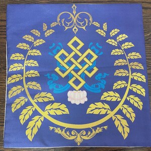 K&ouml;nnte beinhalten: Ein blaues quadratisches Kissen mit einem goldenen Endlosknoten-Design, umgeben von goldenen Bl&auml;ttern und einer wei&szlig;en Blume in der Mitte.