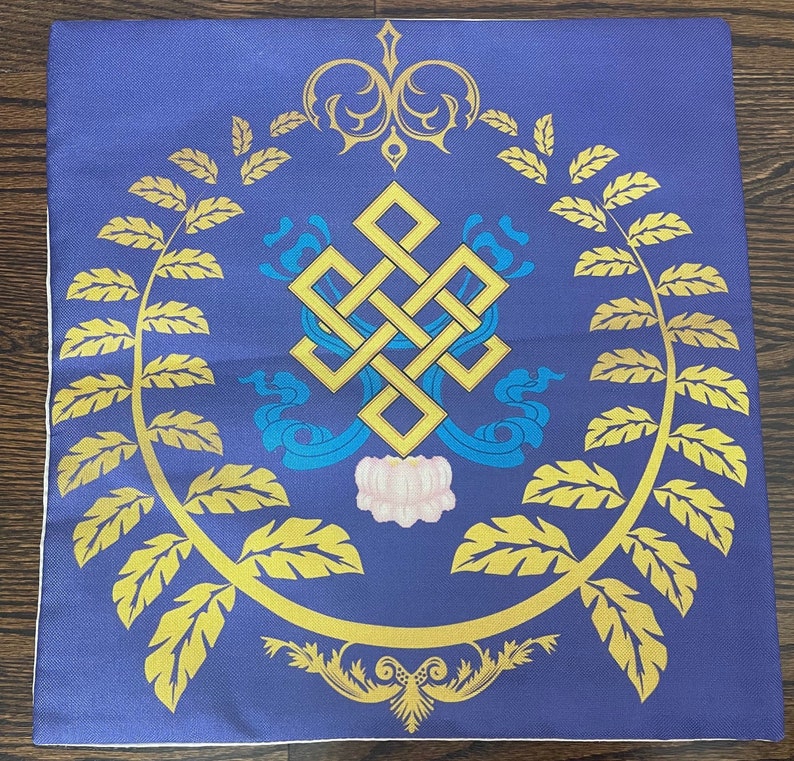 K&ouml;nnte beinhalten: Ein blaues quadratisches Kissen mit einem goldenen Endlosknoten-Design, umgeben von goldenen Bl&auml;ttern und einer wei&szlig;en Blume in der Mitte.