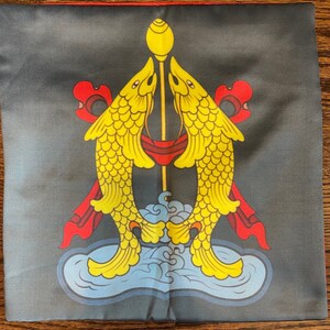 K&ouml;nnte beinhalten: Ein schwarzes Kissen mit einem goldenen und gelben Design aus zwei Fischen, die sich gegen&uuml;berstehen, mit einer goldenen Stange in der Mitte. Die Fische sind von roten und blauen Akzenten umgeben.