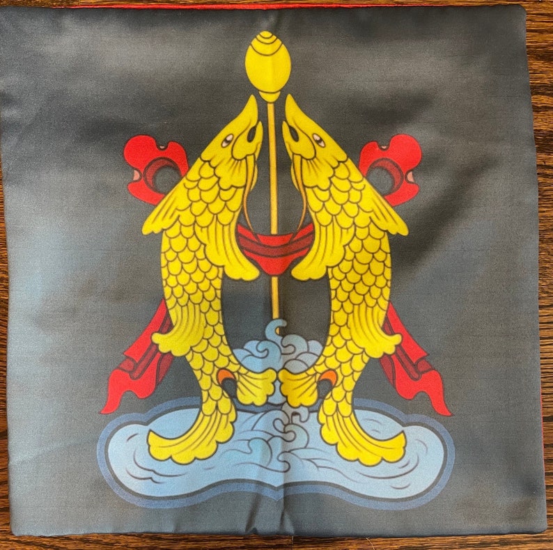 K&ouml;nnte beinhalten: Ein schwarzes Kissen mit einem goldenen und gelben Design aus zwei Fischen, die sich gegen&uuml;berstehen, mit einer goldenen Stange in der Mitte. Die Fische sind von roten und blauen Akzenten umgeben.