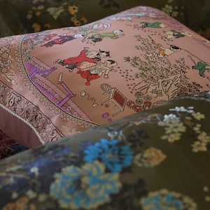 May include: A set of three decorative pillows with floral and children's patterns. The pillows are made of a soft, textured fabric and feature embroidered details. The top pillow has a pink background with a scene of children playing in a garden. The middle pillow has a green background with a floral pattern. The bottom pillow has a green background with a floral pattern.