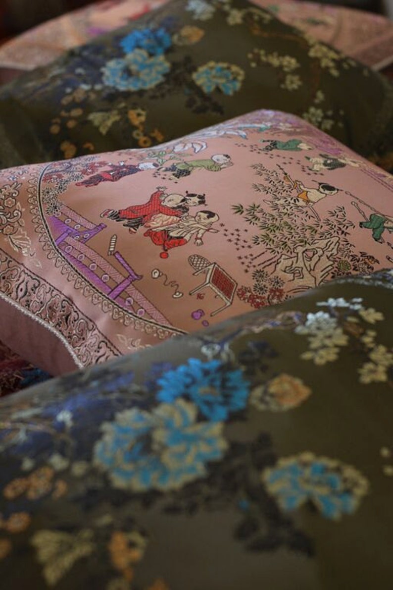 May include: A set of three decorative pillows with floral and children's patterns. The pillows are made of a soft, textured fabric and feature embroidered details. The top pillow has a pink background with a scene of children playing in a garden. The middle pillow has a green background with a floral pattern. The bottom pillow has a green background with a floral pattern.