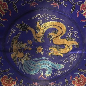 May include: A purple silk cushion with a gold and blue embroidered design of a dragon and a phoenix. The cushion is decorated with floral motifs and clouds.