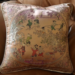 May include: A decorative pillow with a beige background and a gold embroidered border. The pillow features a scene of children playing in a garden with trees and flowers. The children are wearing traditional Chinese clothing.