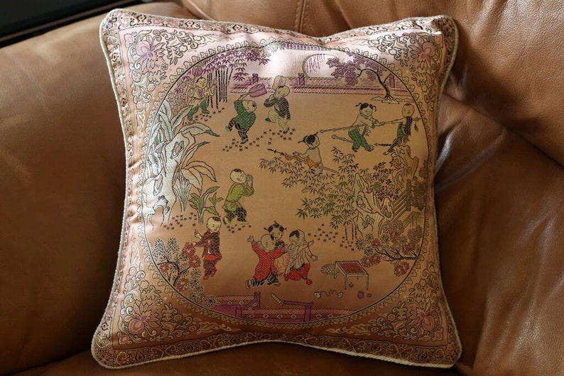 May include: A decorative pillow with a beige background and a gold embroidered border. The pillow features a scene of children playing in a garden with trees and flowers. The children are wearing traditional Chinese clothing.