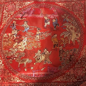 May include: A red square cushion with a gold embroidered border and a circular design of children playing in a garden. The design includes trees, flowers, and a fence.