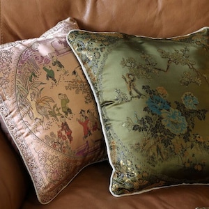 May include: Two decorative throw pillows with embroidered floral and Asian-inspired patterns. The pillow on the left is a light pink with gold accents and the pillow on the right is a dark green with gold accents.