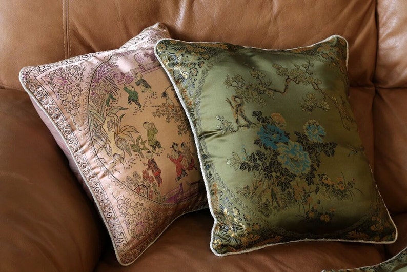 May include: Two decorative throw pillows with embroidered floral and Asian-inspired patterns. The pillow on the left is a light pink with gold accents and the pillow on the right is a dark green with gold accents.