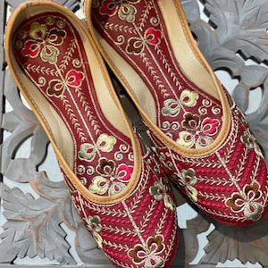 May include: A pair of burgundy slip-on shoes with intricate floral embroidery in gold, pink, and brown. The shoes have a tan leather trim and a soft, cushioned interior. The design features a repeating pattern of embroidered flowers and leaves.