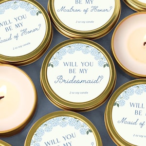 May include: Gold-rimmed soy candles with floral accents and text reading "Will You Be My Bridesmaid?" or "Will You Be My Maid of Honor?" The candles are in small, round tins, some lit, with a soft pink wax.