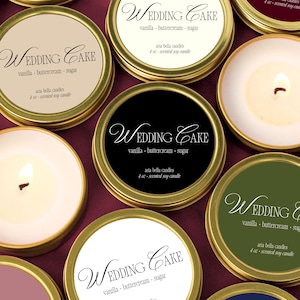 May include: A collection of small, round candles in gold-colored tins. Each tin has a label that reads "Wedding Cake" with scent details. Some candles are lit, showing a warm glow. The background is a deep burgundy.