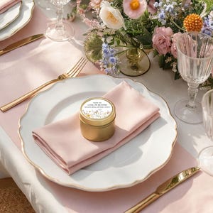 May include: A small gold-colored metal tin with a circular label reading "THANK YOU LOVE IN BLOOM TAYLOR'S BRIDAL SHOWER 2.09.2025" and a floral illustration, resting on a light pink folded napkin atop a white scalloped dinner plate.
