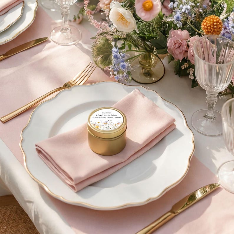 May include: A small gold-colored metal tin with a circular label reading "THANK YOU LOVE IN BLOOM TAYLOR'S BRIDAL SHOWER 2.09.2025" and a floral illustration, resting on a light pink folded napkin atop a white scalloped dinner plate.