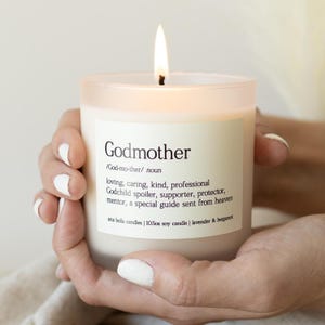 May include: A lit, white soy candle in a frosted glass jar. The candle has a label with the word "Godmother" and its definition. The candle is 10.5oz and scented with lavender and bergamot.