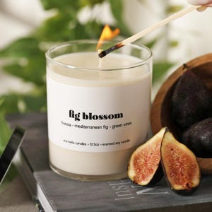 May include: A lit scented soy candle in a clear glass jar labeled "fig blossom." The label lists freesia, mediterranean fig, and green vines. Two fig halves and whole figs are in a wooden bowl. The candle is 10.5 oz.